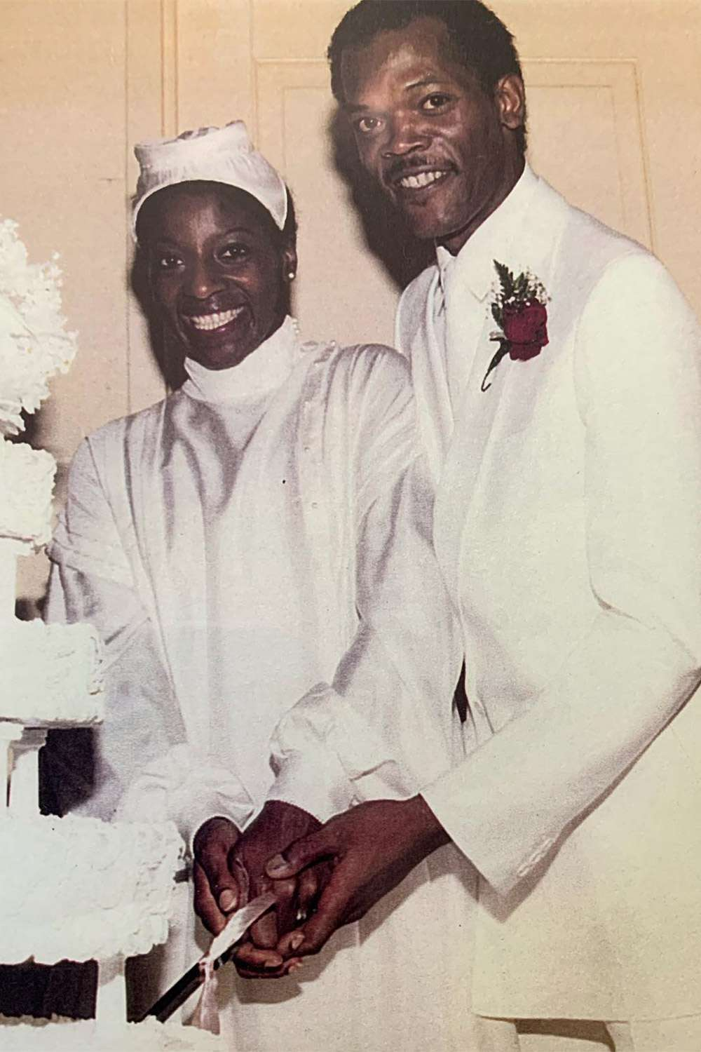 A smiling couple in white outfits cuts a wedding cake together. The woman wears a dress and headpiece, and the man wears a white suit with a red boutonnière. They stand close, holding the knife together.