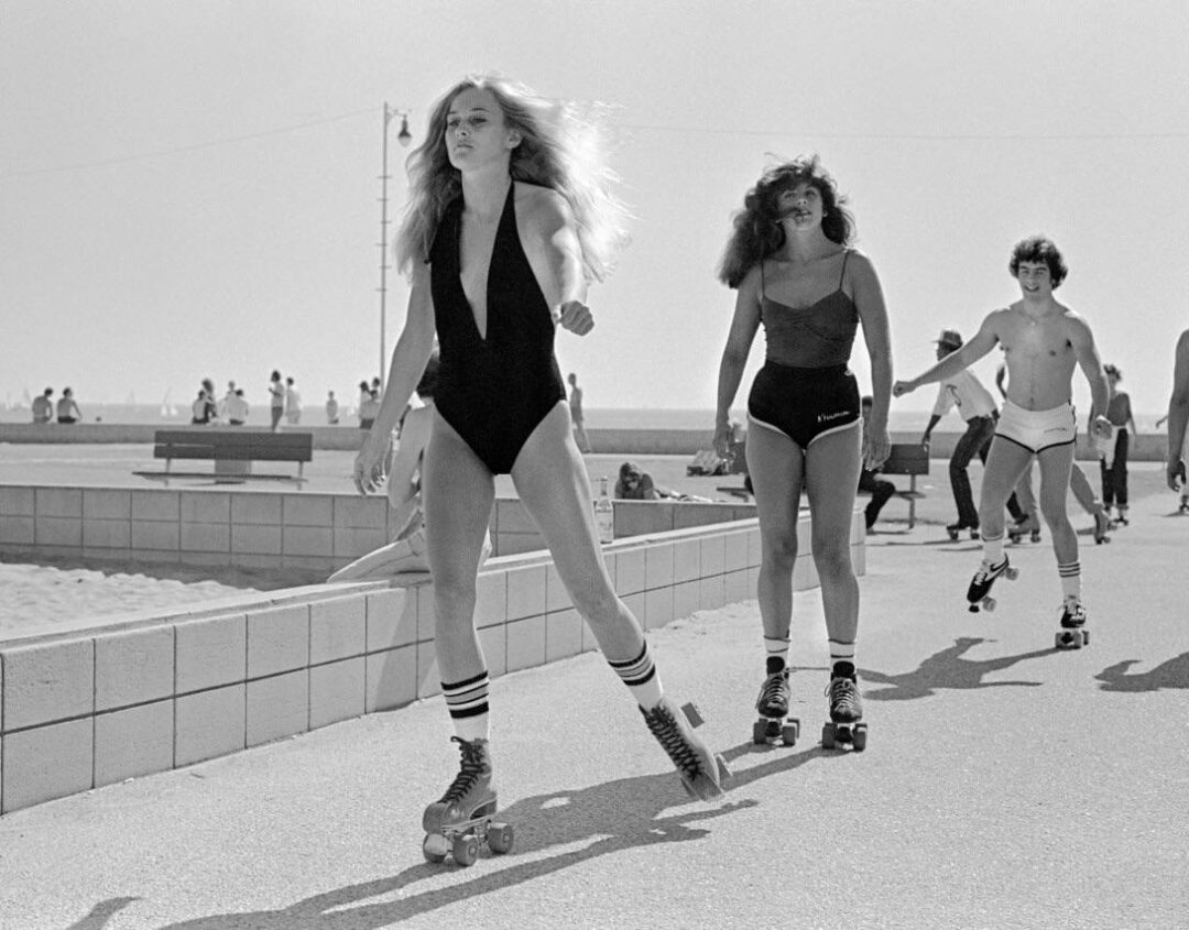 A group of young people roller skating along a paved path near the beach, with two women in swimsuits and striped socks in the foreground. Other skaters and beachgoers are visible in the background.