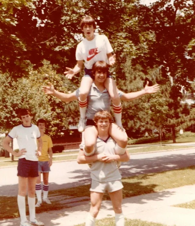 Three young men are stacked on each other's shoulders, smiling and posing outdoors on a sunny day, while two others stand nearby. They wear athletic clothes and knee-high socks, surrounded by trees and a suburban street.
