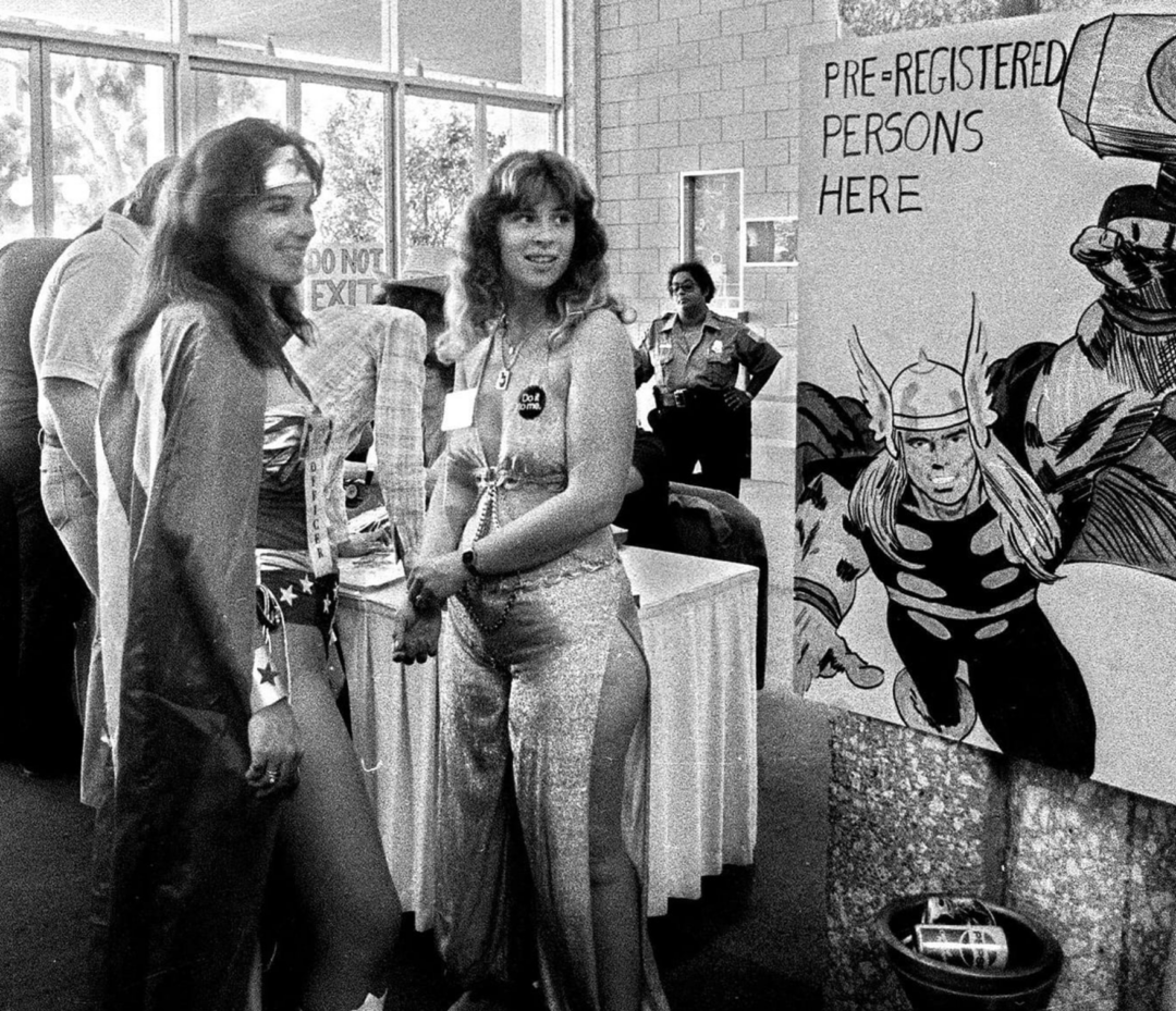Two women in costumes—one as Wonder Woman, the other in a shiny jumpsuit—stand by a table at a convention. A sign reads "Pre-Registered Persons Here" next to a large comic-style drawing of Thor.