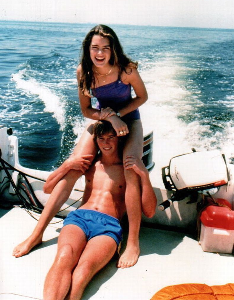 A young woman in a purple swimsuit smiles while sitting on the shoulders of a young man in blue swim trunks; they're on a boat with ocean water in the background on a sunny day.