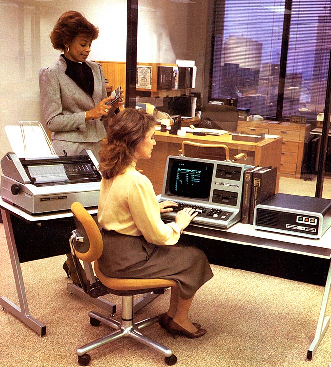 Two women in an office, one seated at a vintage computer typing, the other standing and holding papers. The desk has a large printer, monitor, and other retro computer equipment. Large windows show city buildings outside.