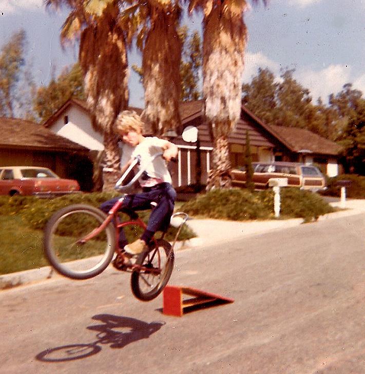 A young boy on a bicycle performs a jump off a small ramp on a suburban street lined with palm trees, houses, and parked cars under a clear sky.