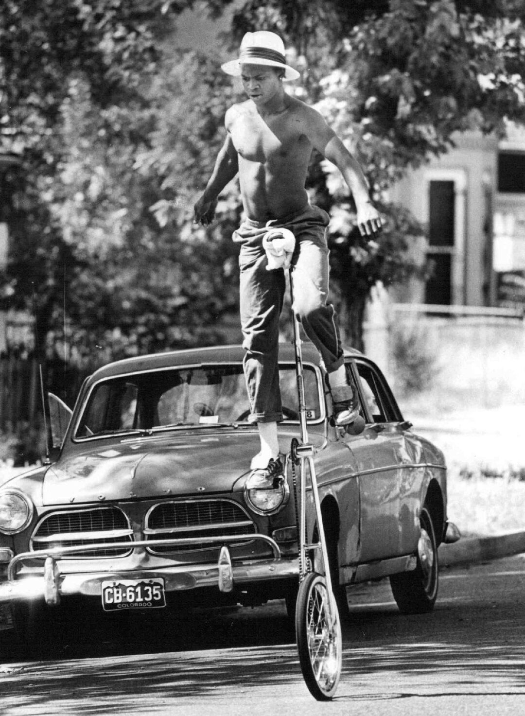 A shirtless man wearing a straw hat and rolled-up jeans rides a tall unicycle on a street, passing in front of a vintage car with trees and houses in the background.