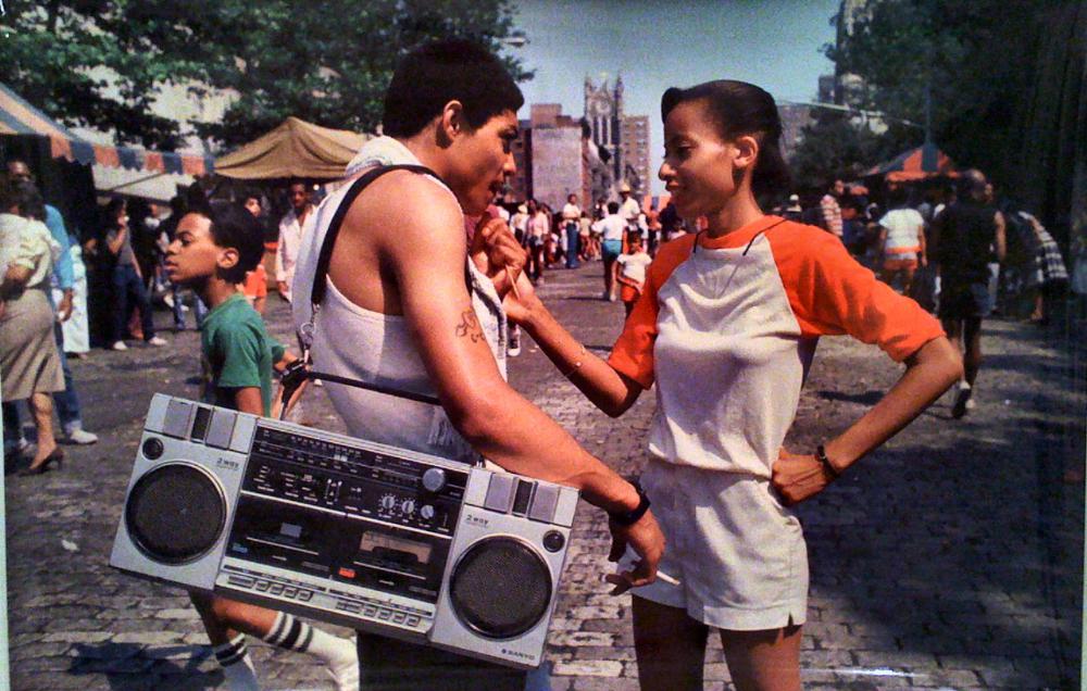 A man with a large boombox on his shoulder talks to a woman in a white and red outfit at an outdoor street market, while people walk by in the background on a cobblestone street.