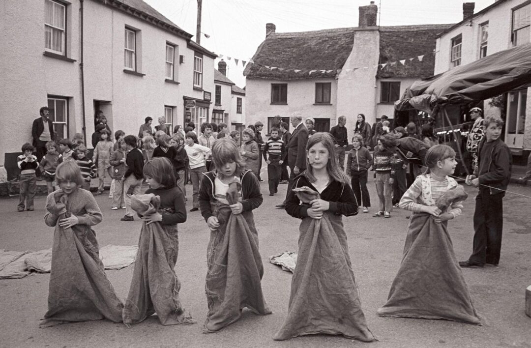 Black-and-white photo of children lined up for a sack race at an outdoor street event, with more children and adults gathered in the background near village houses and bunting overhead.