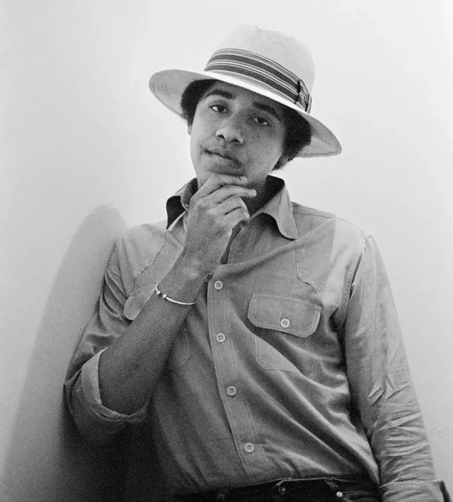 Black-and-white photo of a young man wearing a light-colored hat and button-up shirt, leaning against a wall with his hand thoughtfully resting on his chin.