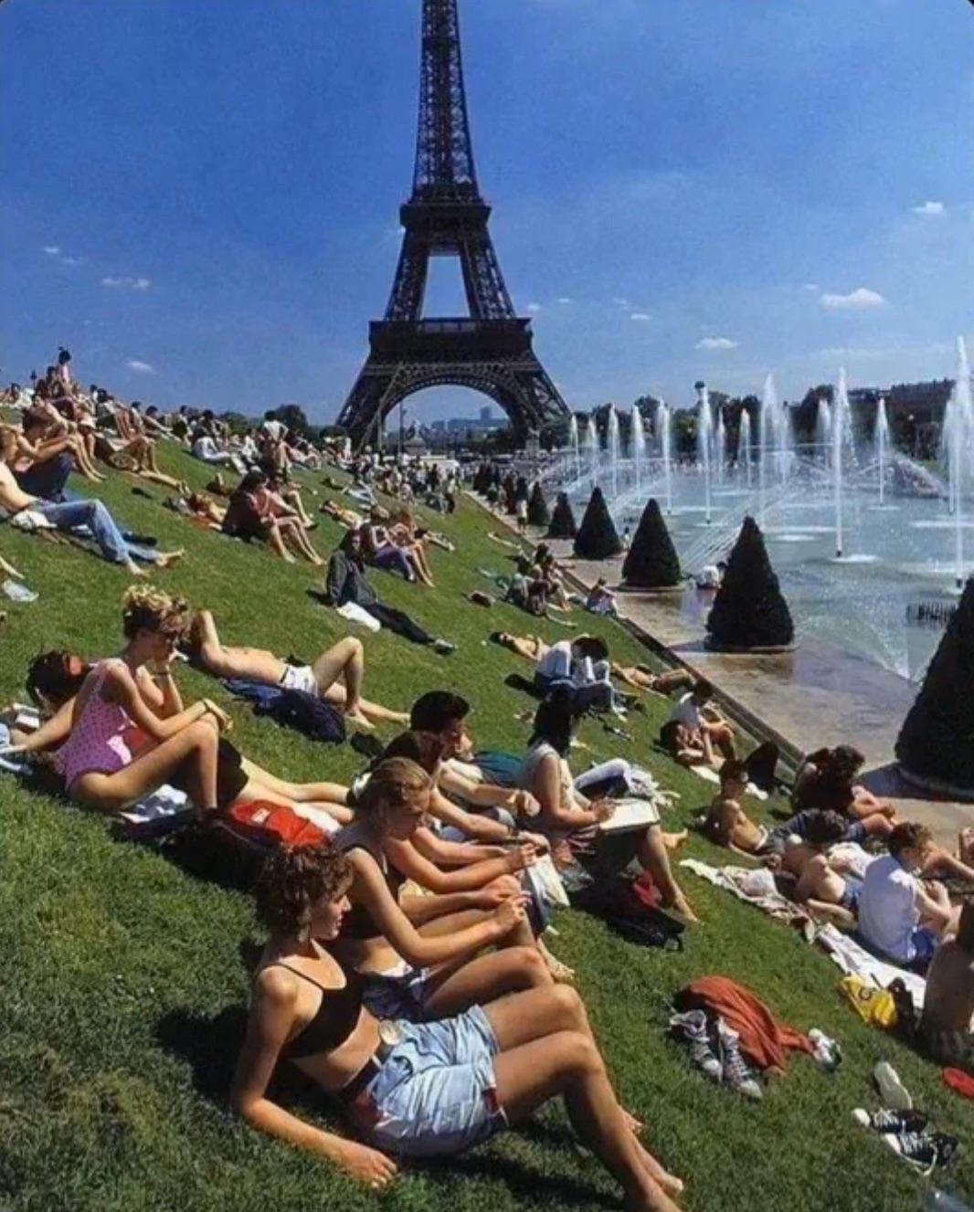 People relax and sunbathe on a grassy slope near fountains by the Eiffel Tower in Paris on a sunny day. The sky is clear and blue, and the area is lively with groups enjoying the outdoors.