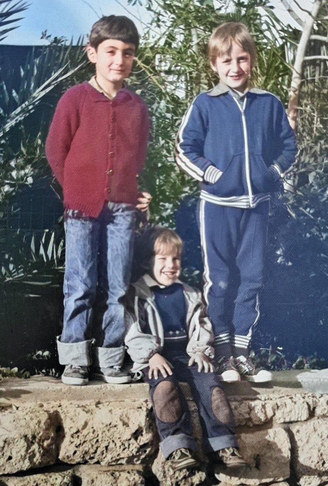 Three children pose outdoors on a stone ledge. Two stand in the back—one wears a red sweater and jeans, the other a blue tracksuit. A third child, wearing a dark jacket, sits in front. Greenery and sunlight are visible in the background.