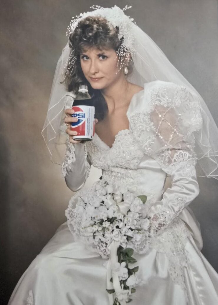 A bride in a white lace wedding dress and veil holds a bouquet of white flowers in one hand and a can of Pepsi in the other, posing for a studio portrait against a neutral background.