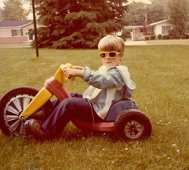 A young child wearing yellow sunglasses and a blue jacket rides a large plastic tricycle on a grassy lawn, with houses, trees, and a basketball hoop visible in the background.