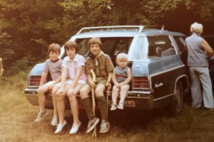 Four children sit on the back bumper of a blue station wagon parked in a grassy area; one child wears a Boy Scout uniform. An adult stands by the driver’s door with their back turned. Trees fill the background.