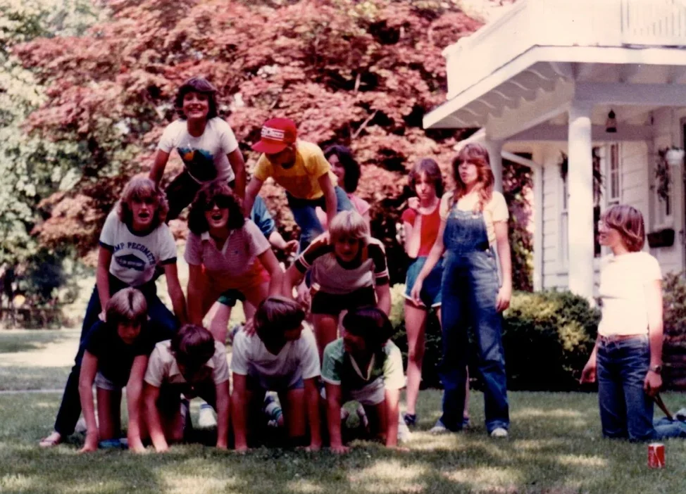 A group of kids form a human pyramid on a grassy lawn, with others standing and watching nearby. Behind them is a large white house and leafy trees on a sunny day.