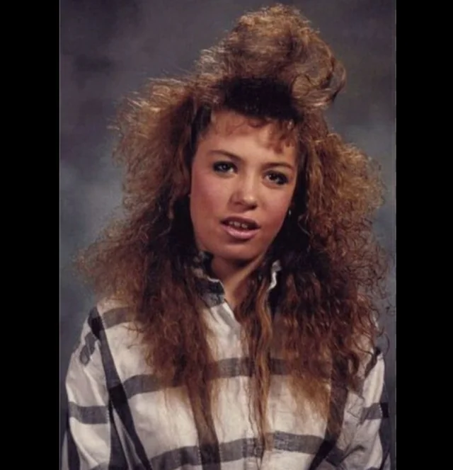 A young woman with voluminous, teased curly hair wears a black-and-white plaid shirt. She stands against a gray studio backdrop, resembling a school portrait from the 1980s or 1990s.