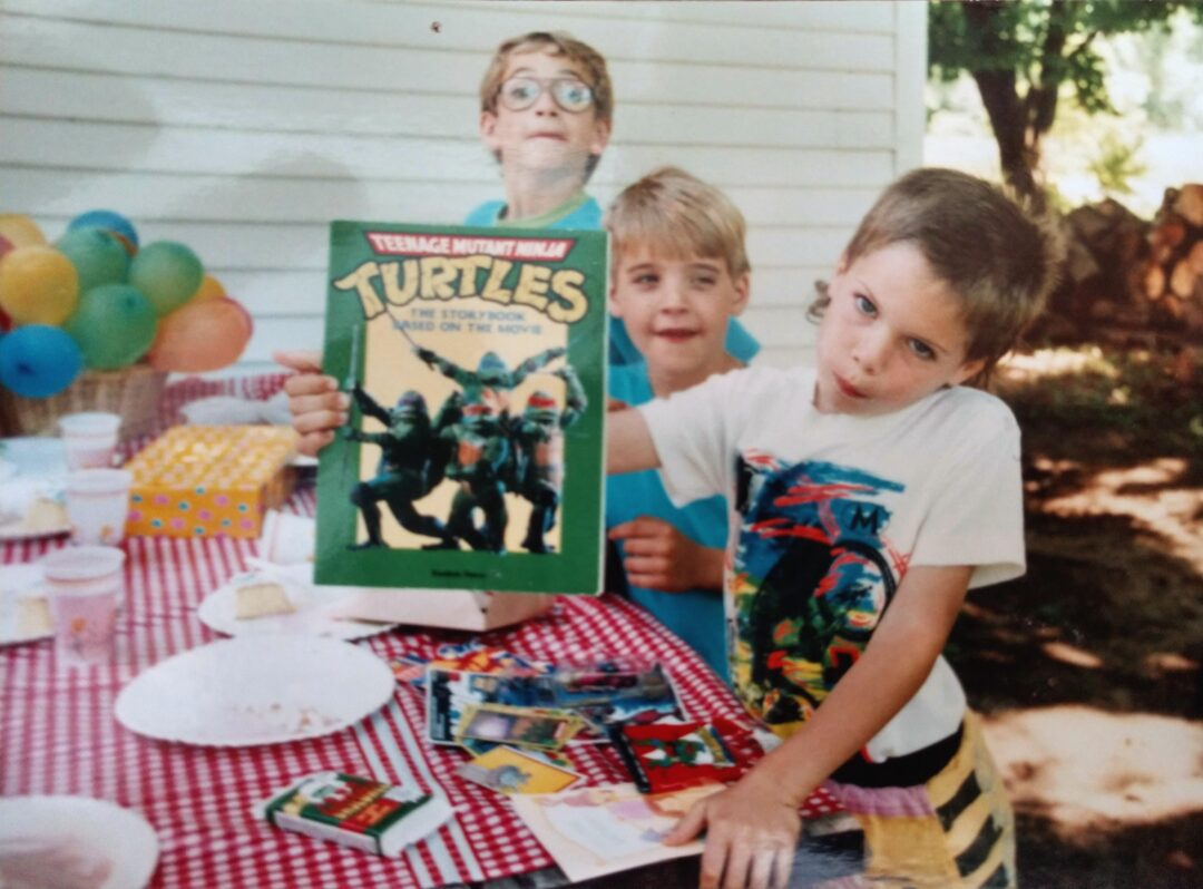 Three young boys at an outdoor party, one holding up a Teenage Mutant Ninja Turtles book. The table has a red checkered cloth, plates, snacks, and colorful balloons in the background.