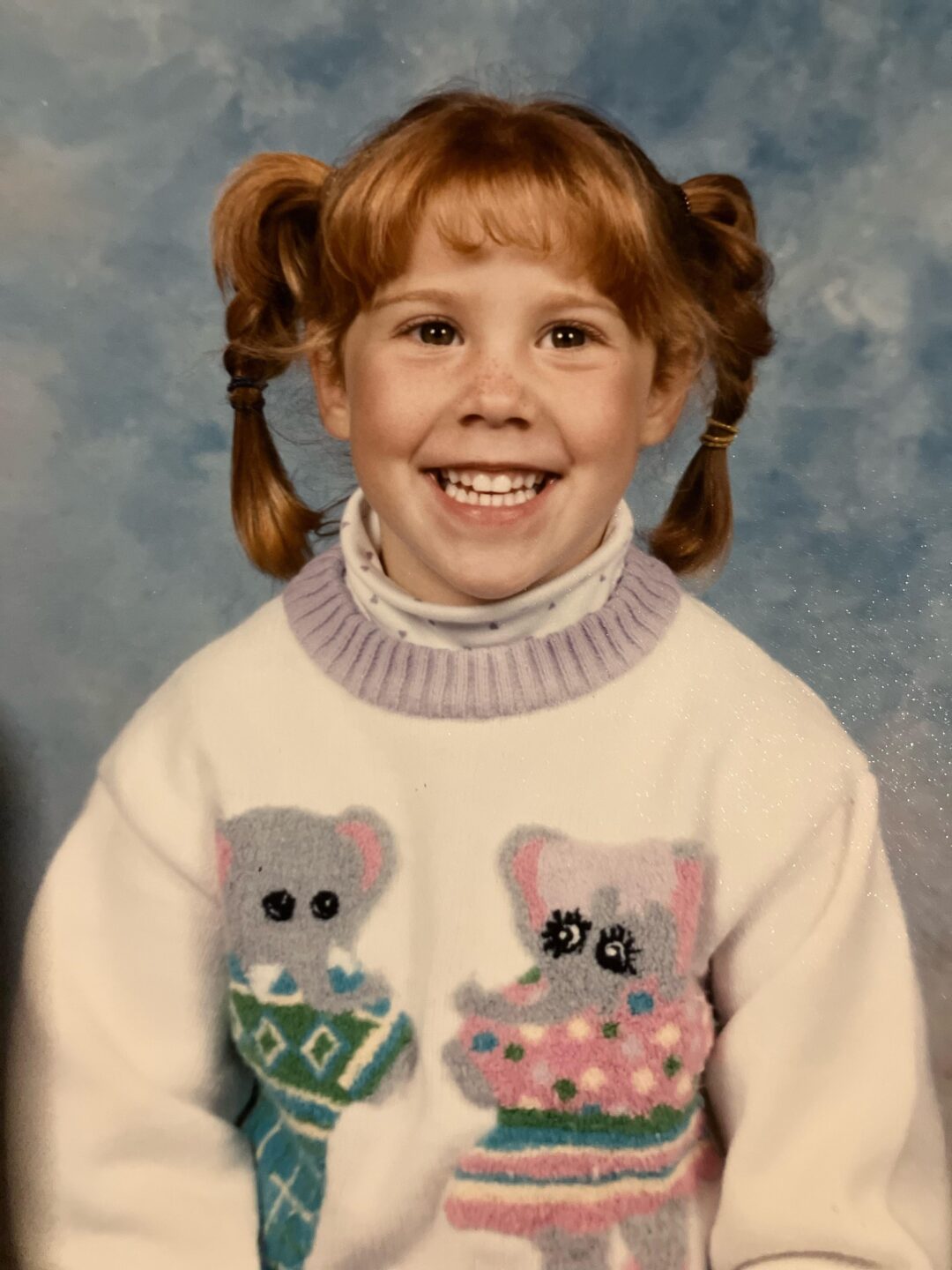 A young girl with red hair in pigtails smiles widely. She wears a white sweater with colorful koalas and a lavender collar, posing in front of a blue mottled studio background.