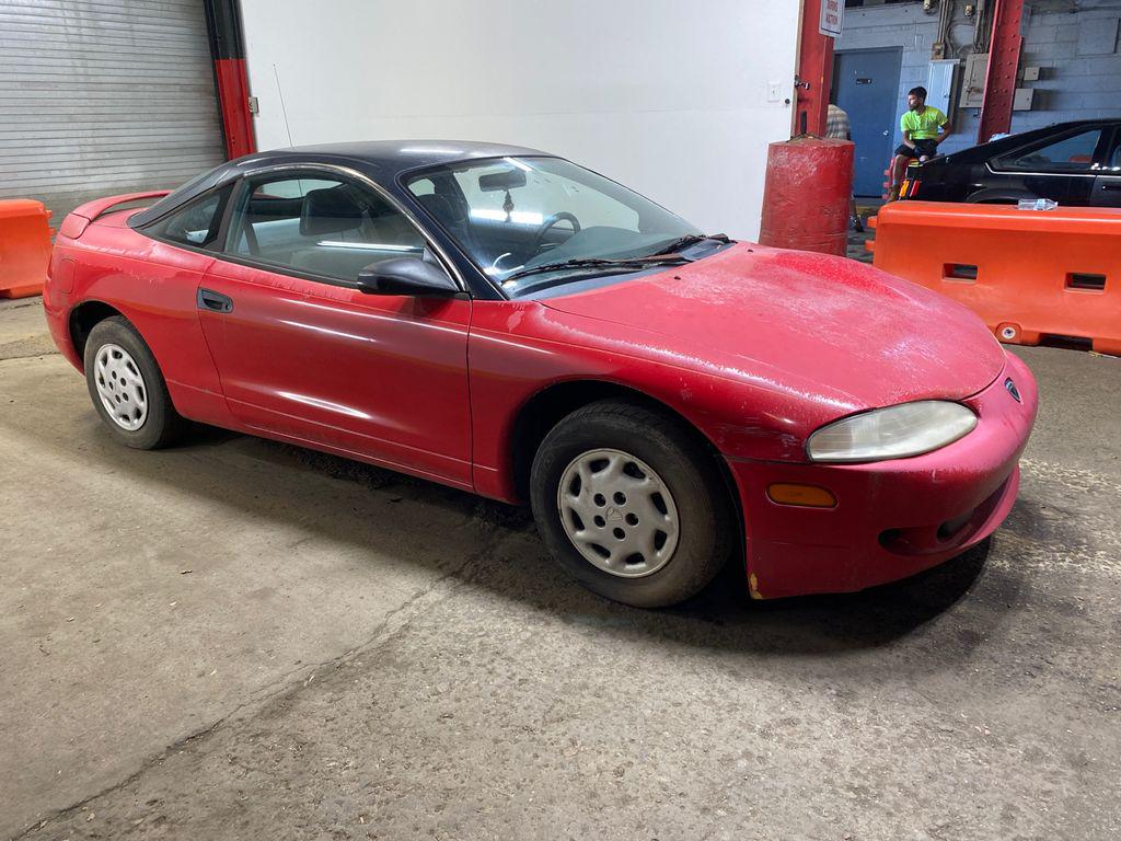 A red two-door coupe with a black roof is parked indoors on a concrete floor near orange barriers and a garage door. The car shows signs of dirt and wear.