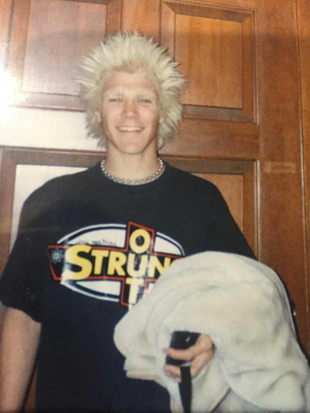 A smiling person with spiky bleached blonde hair stands in front of wooden doors, wearing a black graphic t-shirt and a beaded necklace, holding a light-colored jacket and a strap.