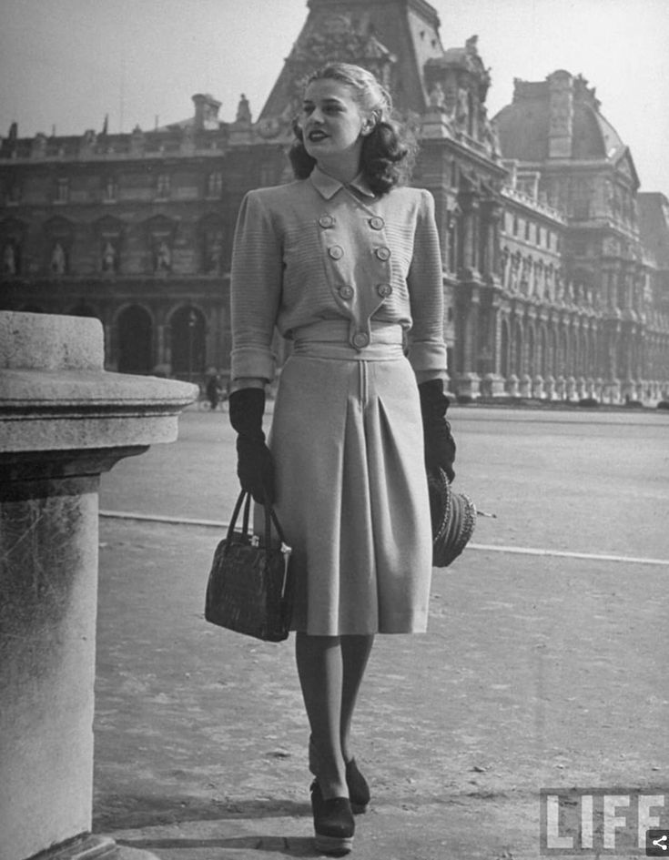 A stylish woman in 1940s fashion, wearing a buttoned jacket, pleated skirt, gloves, and heels, stands outdoors holding a handbag, with a historic building and the LIFE logo visible in the background.