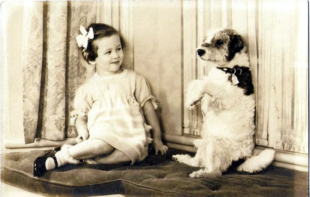 A vintage photo of a young girl in a dress sitting on a cushioned bench, smiling at a small dog that is sitting upright on its hind legs beside her. Both appear relaxed and happy indoors.
