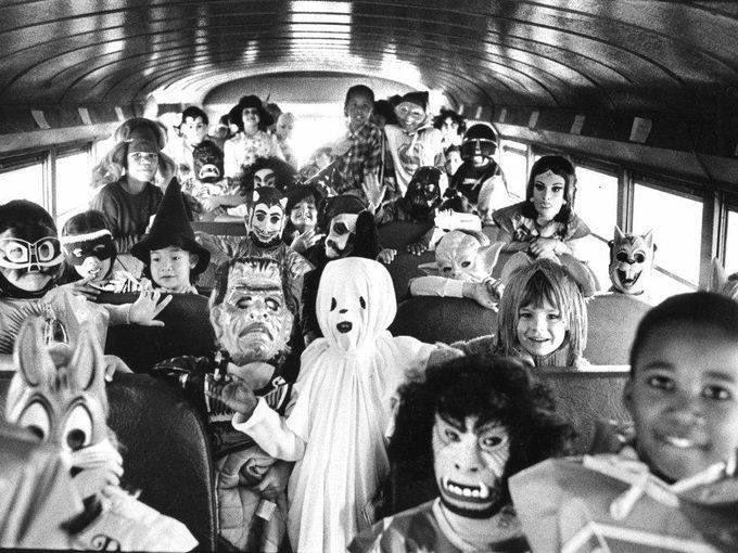 A group of children in Halloween costumes and masks sit on a school bus, smiling and looking at the camera. The costumes include ghosts, witches, animals, and various creatures. The photo is in black and white.