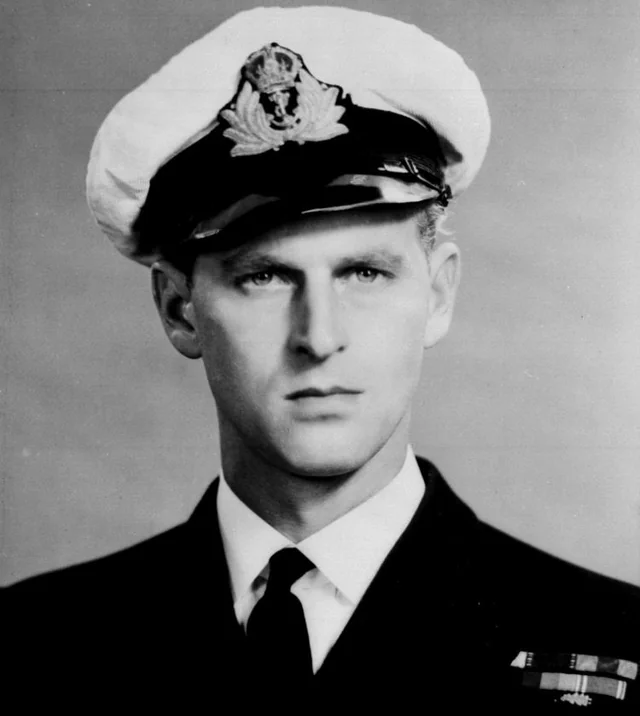 A young man in a formal naval uniform with a peaked cap, displaying an insignia, looks directly at the camera in this black and white portrait. He wears a dark suit and tie, with military ribbons on his chest.