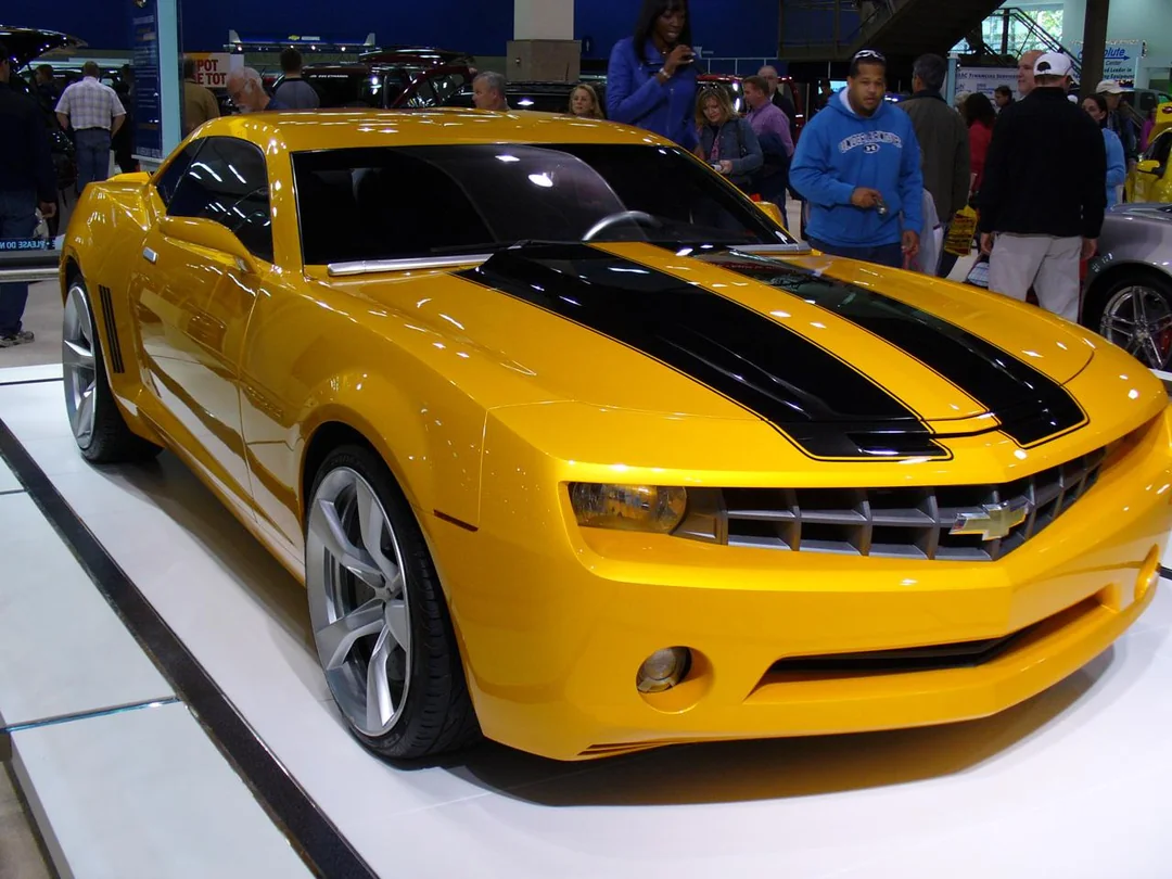 A yellow Chevrolet Camaro sports car with black racing stripes is displayed at an indoor car show, with people standing and admiring it in the background.