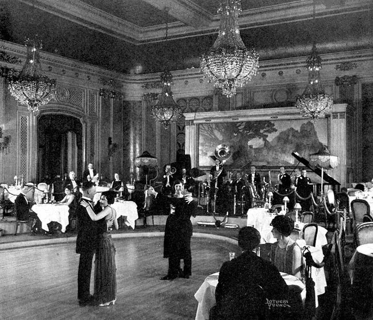 A black and white photo of an elegant ballroom with chandeliers, a live band on stage, and couples in formal attire dancing while others sit at tables around the dance floor.