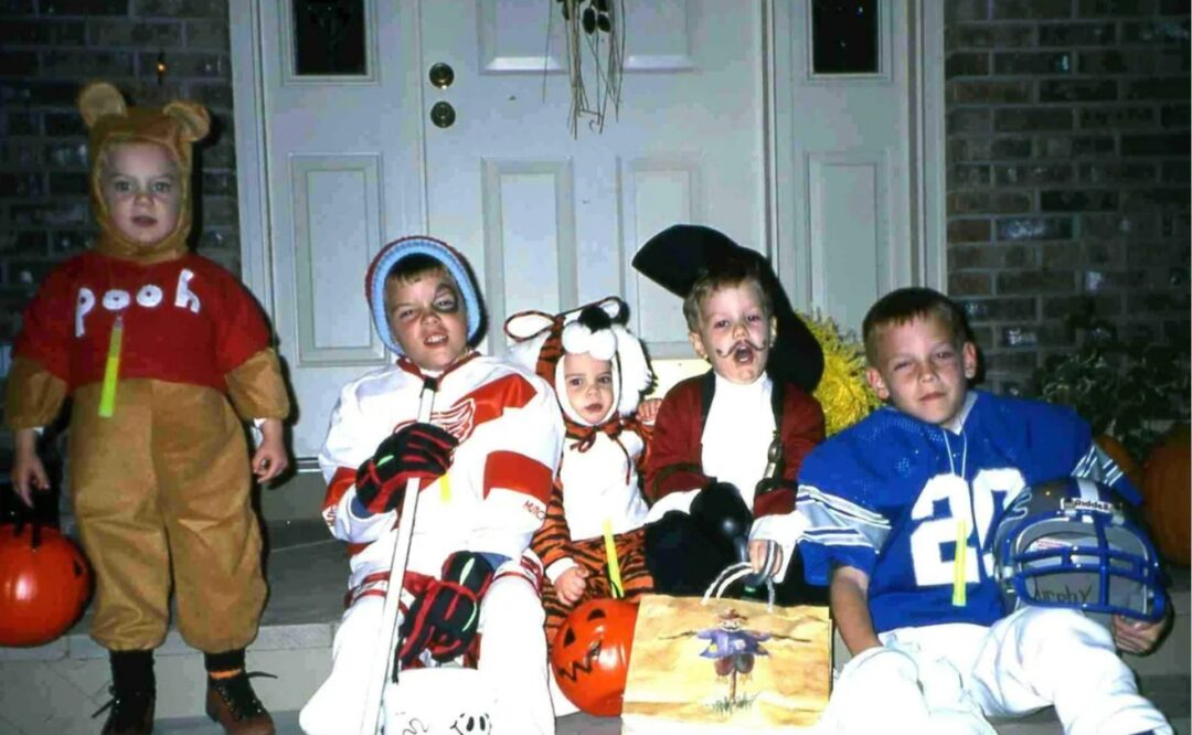 Five children in Halloween costumes—Pooh bear, hockey player, fox, pirate, and football player—sit on a porch with trick-or-treat bags and pumpkins near a front door.