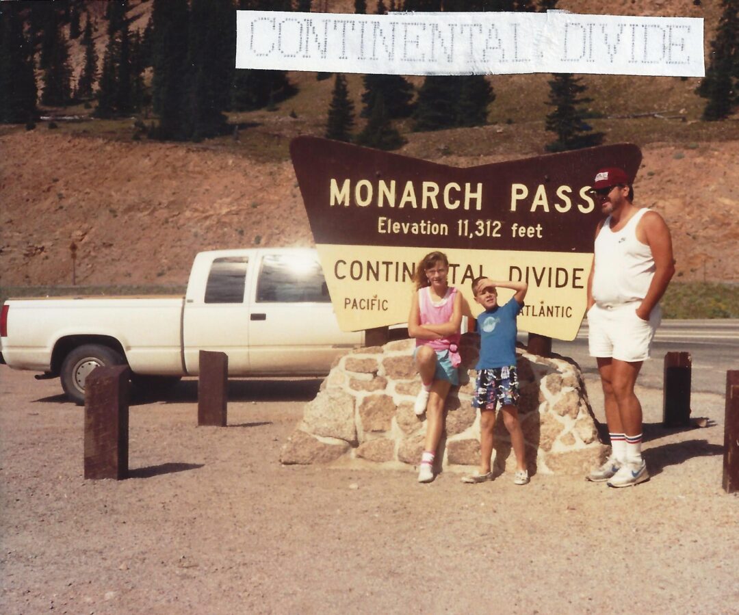 Three people pose in front of a large Monarch Pass sign reading "Elevation 11,312 feet, Continental Divide" with a white pickup truck parked nearby and mountains and trees in the background.
