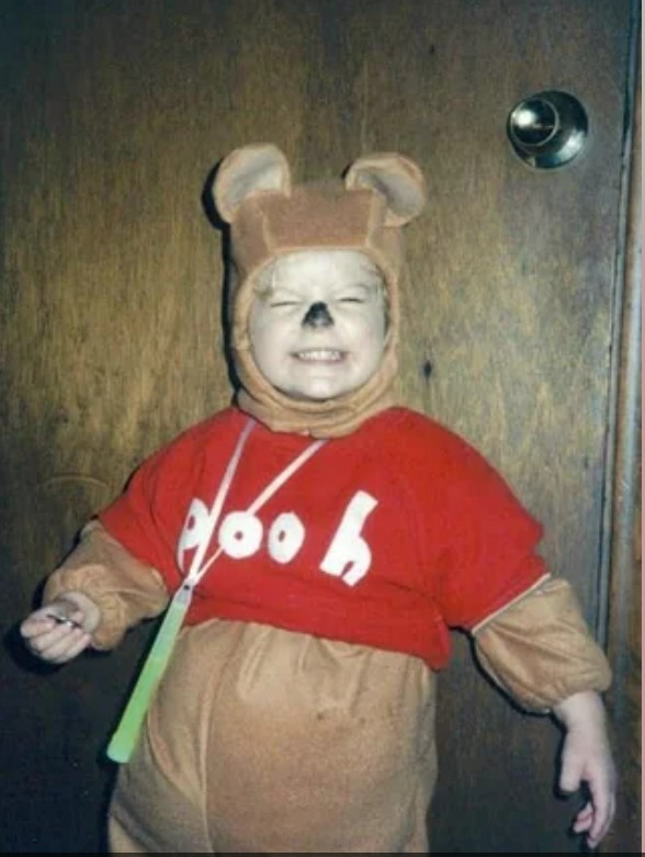 A young child smiles widely while wearing a Winnie the Pooh costume, with bear ears, a red shirt labeled "Pooh," and a painted black nose, standing in front of a wooden door.