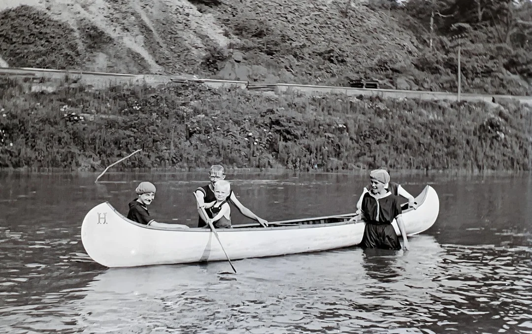 Four children in old-fashioned swimsuits sit and paddle in a white canoe on a calm river, with grassy and hilly terrain visible in the background.