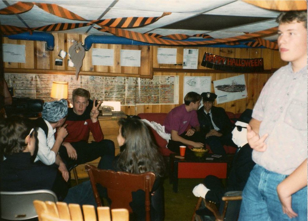 A group of teens in costumes gather in a wood-paneled basement decorated with orange and black streamers and a "Happy Halloween" sign. Some are seated and talking, while one boy stands at the right edge of the photo.