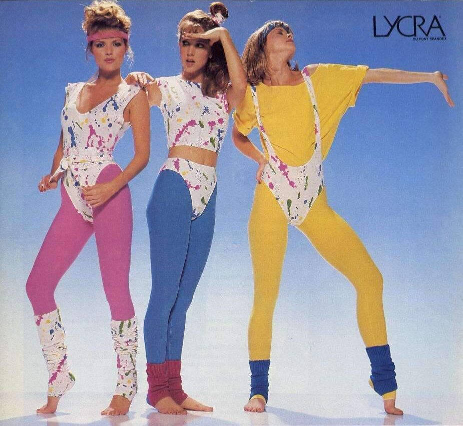 Three women wearing colorful 1980s workout outfits with splatter patterns, leggings, leg warmers, and headbands, pose against a blue background under a "LYCRA" logo.