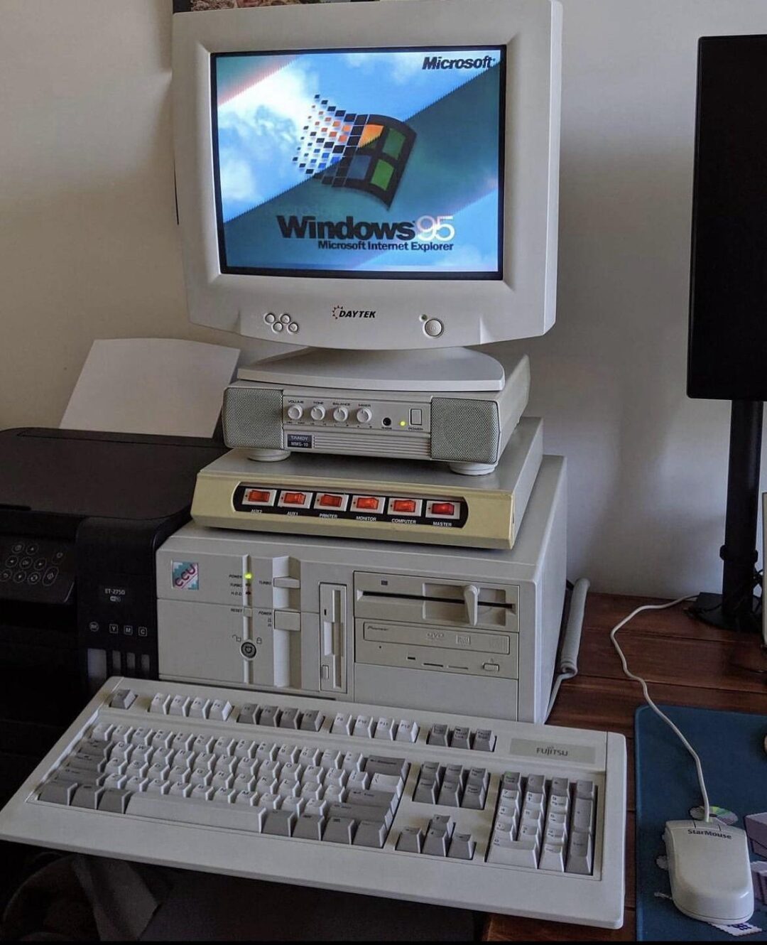 A vintage desktop computer setup with a CRT monitor displaying the Windows 95 startup screen, a beige tower case, floppy disk drives, external speakers, and a classic keyboard and mouse.