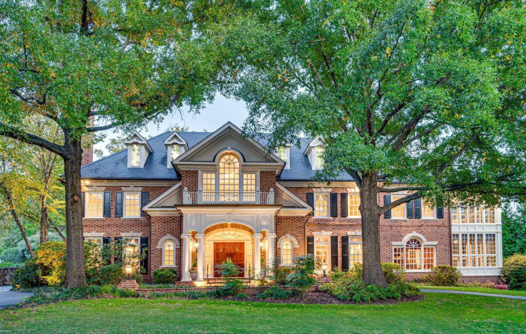 A large, elegant brick house with bright interior lights, arched windows, and decorative columns, surrounded by lush green landscaping and tall trees in the front yard.