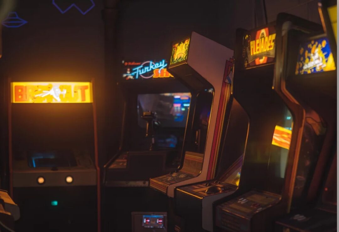 A row of vintage arcade game machines in a dimly lit room, with glowing screens and colorful marquees displaying game titles like "Big Buck" and "Fleance.