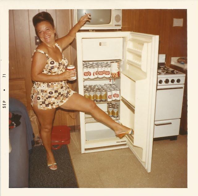 A smiling woman in a patterned swimsuit poses with one leg up on an open fridge stocked with beer cans, holding a can in one hand and gesturing to a small TV on top of the fridge. The scene is in a retro kitchen.