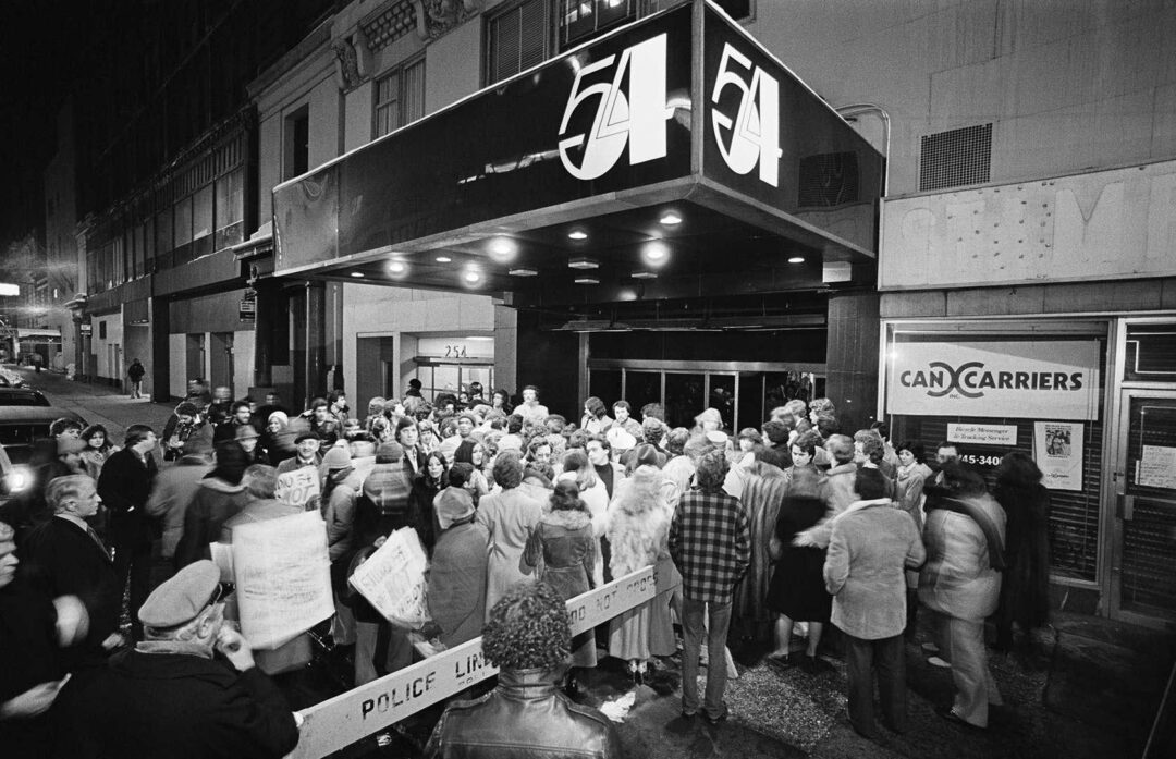 A large crowd gathers outside the entrance of Studio 54 at night, beneath the club’s illuminated sign. Police barricades and a busy sidewalk add to the lively atmosphere.