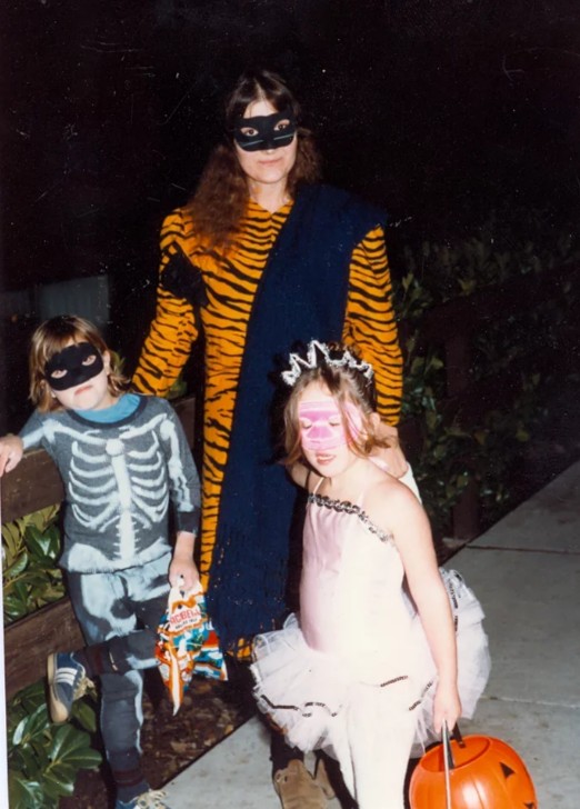 A woman and two children in Halloween costumes stand outside at night. The woman wears a tiger-striped outfit and black mask, the boy is in a skeleton costume, and the girl is a ballerina with a tiara, holding a pumpkin bucket.