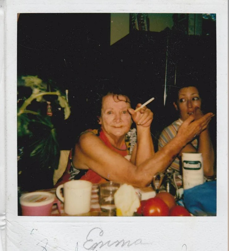 An older woman smiles and gestures with a cigarette at a cluttered table with cups, tomatoes, and containers. Another woman sits beside her, looking to the side. The scene is warm and casual, like a candid moment in a kitchen.