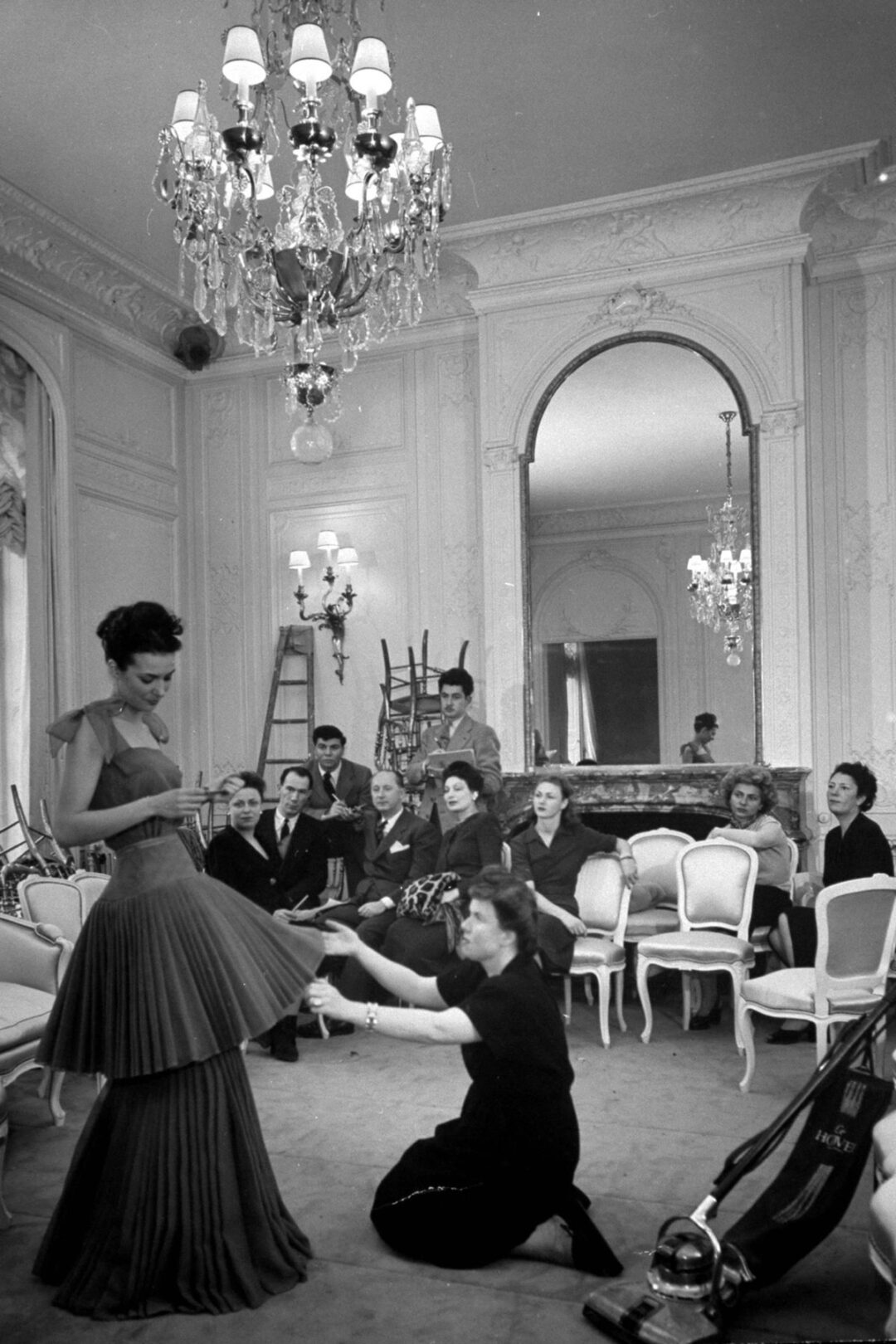 A group of elegantly dressed women watch as a designer adjusts a model’s flowing dress in a luxurious room with chandeliers, ornate mirrors, and white chairs. The scene reflects a classic mid-20th century fashion fitting.