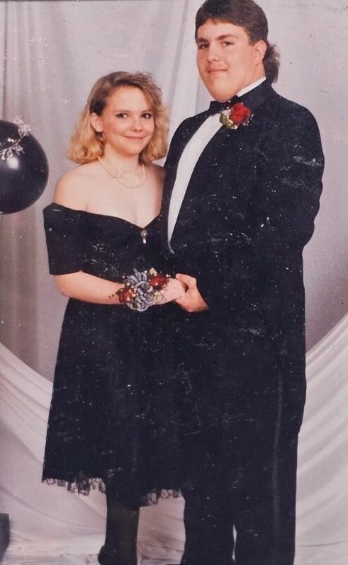 A young couple dressed formally pose for a photo. The woman wears a black off-shoulder dress and corsage, while the man is in a tuxedo with a red boutonniere. They stand together, smiling, against a decorated backdrop.