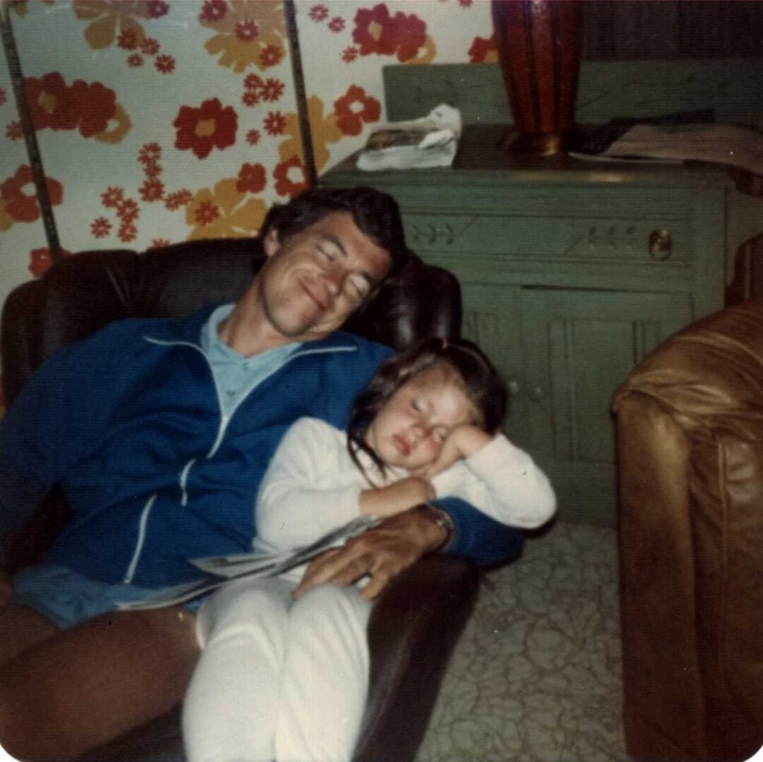 A man and a young girl are asleep together in a brown armchair. The man wears a blue jacket and shorts, and the girl is dressed in white. Behind them is a green cabinet and a wall with bright floral wallpaper.