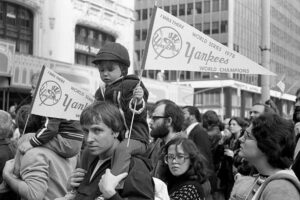 A black-and-white photo of a crowd, including a child on an adult’s shoulders holding a “Yankees World Series 1978 Champions” pennant, during a parade or celebration in a city street.
