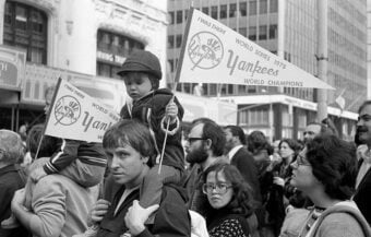 A black-and-white photo of a crowd, including a child on an adult&rsquo;s shoulders holding a &ldquo;Yankees World Series 1978 Champions&rdquo; pennant, during a parade or celebration in a city street.