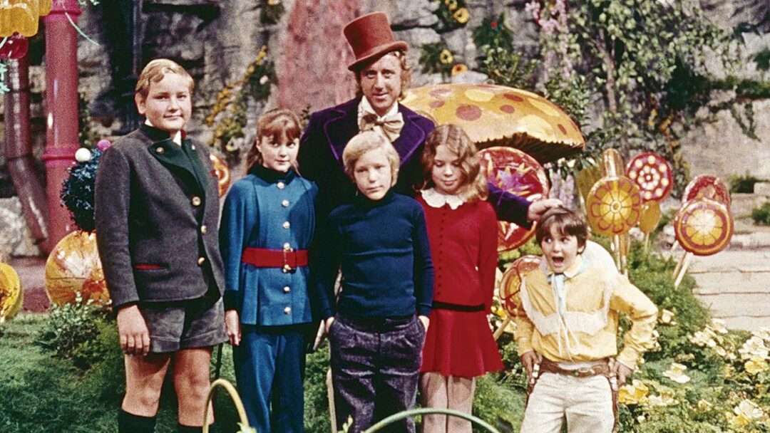 A man in a purple coat and top hat stands with five children in colorful outfits in a whimsical garden filled with oversized candy and plants.