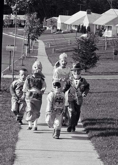 Five children in Halloween costumes, including Batman and other characters, walk excitedly down a suburban sidewalk in a residential neighborhood on a sunny day.