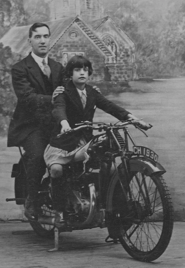 A man in a suit sits behind a young girl on a vintage motorcycle with the license plate "CH1930." They pose in front of a painted backdrop of a building and trees, in a black-and-white, early 20th-century photograph.