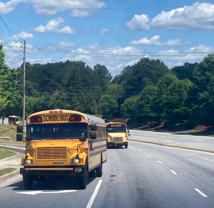 Two yellow school buses drive down a tree-lined road under a blue sky with scattered clouds. The road is wide and mostly empty, with one bus closer to the foreground and another in the distance.